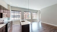 This single-floor unit offers a thoughtfully designed open-concept layout that seamlessly connects the kitchen and living areas. The kitchen serves as the centerpiece of the space, anchored by a large dark-stained island with a white quartz countertop and an undermount sink. Matching dark cabinetry extends to ceiling height, providing ample storage, while stainless steel appliances — including a refrigerator, electric range, and microwave — complete the modern setup. Globe pendant lights hang above the island, adding a stylish, contemporary touch. Oversized black-framed windows flood the open living area with natural light and offer views of the surrounding urban streetscape, contributing to an airy, loft-inspired ambiance. Rich hardwood-style flooring runs throughout the entire unit, providing warmth and visual continuity. The bedroom is generously proportioned, featuring neutral-toned walls and the same hardwood flooring, creating a calm and versatile space. The bathroom is well-appointed with a dark-stained vanity, white quartz countertop, a large mirror, and a full soaking tub with a subway-tiled surround. The overall design emphasizes clean lines, neutral tones, and modern finishes throughout.