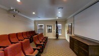 This dedicated home theater room delivers an authentic cinema experience within a single-floor layout. The space is outfitted with rows of traditional stadium-style seating upholstered in rich red fabric, complete with fold-down cushions and armrests — closely mirroring the feel of a classic movie house. The seats are arranged to provide clear sightlines to a large projection screen mounted along one wall, ensuring an unobstructed view from every position. A dark wood media console sits adjacent to the screen, offering storage and display functionality. The room is finished with warm neutral-toned walls accented by elegant crown molding, lending a refined, polished character to the space. Recessed lighting in the ceiling provides controlled illumination, ideal for setting the mood before and after screenings. A patterned area rug in earthy tones covers the floor, adding both visual warmth and acoustic comfort. Windows dressed with slatted blinds allow for natural light when desired, while also offering full light control for optimal viewing conditions. A decorative sculptural piece displayed on a pedestal near the entrance adds an artistic focal point, elevating the overall ambiance of this thoughtfully designed entertainment space.