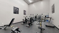 Located at 2881 Richmond Rd in Ottawa, Ontario, this fitness facility offers a focused and functional workout environment within a single, efficiently utilized space. The room features notably high ceilings that lend an open, airy feel despite the compact footprint, with overhead fluorescent lighting ensuring the area is consistently well-lit throughout.

The facility is outfitted with a comprehensive range of cardio equipment, including multiple air bikes, a Star Trac treadmill, a rowing machine, and additional cycling machines. A multi-station strength training unit and cable machine provide options for resistance-based workouts, rounding out the gym's capabilities. Rubber flooring throughout offers durability and safety underfoot.

A large wall-mounted mirror along one side of the room expands the sense of space and allows users to monitor their form during exercise. Two industrial wall-mounted fans provide ventilation and airflow throughout the space. The neutral grey walls serve as a clean backdrop for a series of bold black-and-white athletic photography prints, adding a motivational and professional aesthetic to the environment.

The overall ambiance is sleek and purpose-driven, designed to support focused training sessions in a no-frills, performance-oriented setting.