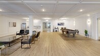 Located at 175 Quincy Ct in Perth Amboy, NJ, this expansive single-floor living space offers a sophisticated and versatile environment designed for both relaxation and recreation. The interior is defined by striking coffered ceilings and crisp white wainscoting panels that lend a refined, classic character throughout.

The open layout is thoughtfully organized into multiple distinct zones. A generously sized lounge area anchors one section of the space, furnished with plush leather sofas and accent chairs arranged around a sleek glass coffee table, creating an inviting setting for socializing. Natural light filters in through large windows dressed with layered sheer and taupe draping curtains, enhancing the warm, airy ambiance.

A dedicated game area features a full-sized billiards table set atop light-toned hardwood flooring that runs seamlessly across the entire floor. Adjacent to this is a card and game table surrounded by upholstered chairs, ideal for more intimate gatherings.

Structural columns with decorative moldings add architectural interest while subtly defining different functional zones. A compact kitchenette with white cabinetry and stainless steel appliances provides convenient amenities. The overall palette of soft whites, warm neutrals, and natural wood tones creates a cohesive, elegant atmosphere suited for community use.