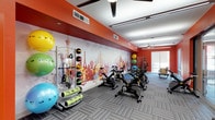 This dynamic fitness studio is designed to inspire an active lifestyle through thoughtful layout and energetic design. The space is anchored by a striking, colorful cityscape mural that spans the length of one wall, featuring iconic landmarks rendered in bold reds, yellows, and whites — creating a lively, motivating atmosphere for workouts.

The studio is outfitted with a row of stationary spin bikes positioned along the central floor space, ideal for group cycling sessions or individual cardio training. Along the walls, a well-organized equipment station offers an array of exercise tools, including medicine balls, resistance bands, foam rollers, and stability balls in a variety of sizes and colors. A dedicated equipment rack keeps everything neatly arranged and accessible.

Overhead, ceiling fans provide airflow throughout the space, complemented by recessed lighting and modern linear pendant lights that ensure even, bright illumination. The flooring consists of durable, patterned carpet tiles in a neutral gray tone, offering both comfort and practicality for a variety of exercise activities.

Natural light filters in through a window at the far end of the room, adding an open, airy quality to the space. The bold orange accent walls tie the room together, creating an energetic and cohesive environment well-suited for a range of fitness activities.
