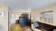 This single-floor studio in Arlington offers a thoughtfully arranged open-concept layout that maximizes its living space. The room is defined by warm hardwood flooring that runs throughout, providing a cohesive and inviting foundation. A distinctive dark wood slatted room divider serves as the primary architectural element separating the sleeping area from the living space, offering privacy while maintaining an open, airy feel.

The sleeping area is tucked behind the divider and features a framed headboard, coordinating nightstands with table lamps, and a window dressed with patterned curtains that allow for light control. A ceiling fan with an integrated light fixture is centrally positioned overhead, ensuring comfort and airflow throughout the space.

The living area accommodates a neutral-toned sofa accented with blue throw pillows and is complemented by a decorative wall piece that adds a touch of character. A textured area rug anchors the living zone. The textured popcorn ceiling and soft, warm wall tones contribute to a cozy and casual ambiance throughout this efficiently designed single-level space.