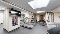 Located at 2112 New Hampshire Ave NW in Washington, DC, this fitness center offers a comprehensive workout facility spread across a single floor. The space is thoughtfully organized into distinct exercise zones, making efficient use of its layout to accommodate a wide variety of training needs.

The cardio area features multiple Star Trac treadmills and an elliptical trainer, complemented by a wall-mounted flat-screen television for entertainment during workouts. Full-length mirrors line portions of the walls, enhancing the sense of openness throughout. A stretching guide display is conveniently positioned nearby for reference.

The strength training section is well-appointed, housing a multi-function cable machine, a Body-Solid power rack with weight plates, and a dedicated free weight area with a multi-tier dumbbell rack. Additional equipment includes a seated rowing machine, a stability ball, and a balance ball, supporting a range of functional fitness routines.

The facility is finished with durable dark rubber flooring throughout, providing both safety and comfort during exercise. Recessed lighting and skylights bring a bright, clean atmosphere to the space, while the neutral cream-toned walls contribute to a focused and professional environment. The fitness center is accessible via a dedicated entry door and is well-suited to serve the residents of the building.