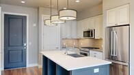 This 2-bedroom, 3-bathroom residence is designed with a modern yet refined aesthetic, offering an efficient and comfortable layout across a single floor. The open-concept kitchen serves as the heart of the home, showcasing a large center island with a white quartz countertop and contrasting dark cabinetry base, a stainless steel appliance suite including a gas range, built-in microwave, and French door refrigerator, and warm tile backsplash beneath classic white raised-panel cabinetry. Three stylish drum pendant lights hang above the island, adding a contemporary focal point. The kitchen flows naturally toward a living area with large sliding glass doors that invite abundant natural light and provide direct access to a private balcony with urban views. Both bedrooms feature durable hardwood-style flooring and neutral tones. The primary bedroom is a comfortable size, benefiting from its own en-suite bathroom with a quartz vanity and tile flooring, along with a dedicated walk-in closet with dual hanging rods and shelving. The second bedroom includes a bright window with exterior views. A dedicated laundry room houses a stacked Whirlpool washer and dryer alongside built-in cabinetry with a folding surface, adding everyday convenience to this well-rounded residence.