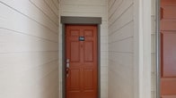 This well-appointed 2-bedroom, 2-bathroom residence offers a thoughtfully designed single-floor layout that maximizes comfort and functionality. The entry greets you with a bold terracotta-colored front door, adding a warm and inviting touch to the exterior.

Inside, the open-concept living area is finished with plush carpeting and neutral tones throughout, creating a bright and welcoming atmosphere. Crown molding accents the ceilings, adding a refined architectural detail to the main living space. The kitchen is seamlessly integrated into the layout via a peninsula with a breakfast bar, framed by a decorative column surround. Dark wood cabinetry, stone countertops, hardwood-style flooring, and stainless steel appliances give the kitchen a polished, modern feel.

The floor plan flows naturally between the living, dining, and kitchen areas, offering generous room for everyday living. Multiple doors visible from the main area lead to the bedrooms and additional closet or storage spaces, suggesting a well-organized and practical layout.

A private balcony extends the living space outdoors, offering open, expansive views of the surrounding landscape under wide open skies — ideal for enjoying morning coffee or evening relaxation. This residence combines comfortable interiors with a connected, airy feel throughout.