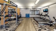 This spacious fitness center is designed to accommodate a full spectrum of workout routines, offering a well-organized and thoughtfully laid-out environment for exercise. The flooring combines durable hardwood-style planks with patterned area rugs, defining distinct workout zones throughout the space. Multiple ceiling fans and overhead fluorescent lighting ensure the facility remains well-ventilated and brightly lit at all times.

The cardio section is generously outfitted with True treadmills, elliptical trainers, a stair climber, and a rowing machine, all positioned near a wall-mounted television for entertainment during workouts. Adjacent to the cardio area, a dedicated strength training zone features a multi-station cable and weight machine, a Smith machine with plate-loaded barbells, a full dumbbell rack spanning a wide range of weights, and a cable row machine.

A large open floor area in the center of the space, accented by an orange accent wall, provides ample room for stretching, mat exercises, or functional training, complemented by medicine balls and a folded exercise mat stored nearby. Floor-to-ceiling mirrors line portions of the walls, enhancing the sense of openness. Glass double doors open to an outdoor area, allowing natural light to filter in and providing easy access to the exterior. The overall ambiance is clean, modern, and purposefully designed for an effective fitness experience.