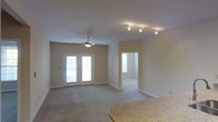 This single-story space presents an open, functional layout centered around a spacious living area finished with neutral-toned walls and light carpeting that contributes to a clean, understated aesthetic. The living area is illuminated by a combination of a ceiling fan with an integrated light fixture and a modern track lighting system mounted on the ceiling, offering versatile lighting options throughout the day. A set of white-framed French doors provides direct access to the outdoors, allowing natural light to filter into the room and creating a welcoming sense of openness.

Adjacent to the living area, an arched or squared doorway leads into a secondary room, also benefiting from its own window that draws in additional natural light. This connected layout enhances the overall flow of the space. In the foreground, a granite-style countertop with a curved edge suggests a nearby kitchen or bar area, indicating that the living and dining or kitchen spaces share an integrated relationship. The white baseboards throughout provide a crisp contrast to the neutral walls, while the overall design remains practical and move-in ready.