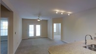 This single-story space presents an open, functional layout centered around a spacious living area finished with neutral-toned walls and light carpeting that contributes to a clean, understated aesthetic. The living area is illuminated by a combination of a ceiling fan with an integrated light fixture and a modern track lighting system mounted on the ceiling, offering versatile lighting options throughout the day. A set of white-framed French doors provides direct access to the outdoors, allowing natural light to filter into the room and creating a welcoming sense of openness.

Adjacent to the living area, an arched or squared doorway leads into a secondary room, also benefiting from its own window that draws in additional natural light. This connected layout enhances the overall flow of the space. In the foreground, a granite-style countertop with a curved edge suggests a nearby kitchen or bar area, indicating that the living and dining or kitchen spaces share an integrated relationship. The white baseboards throughout provide a crisp contrast to the neutral walls, while the overall design remains practical and move-in ready.
