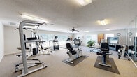 This fitness center offers a generously sized, single-floor workout space designed to accommodate a wide variety of exercise needs. The open layout is bright and welcoming, with multiple windows lining the walls and allowing abundant natural light to fill the room, creating an energizing atmosphere for workouts.

The space is outfitted with an extensive selection of True-brand cardio equipment, including upright and recumbent stationary bikes, treadmills, and elliptical machines, all strategically positioned on protective floor mats throughout the room. A multi-station cable weight machine anchors the strength training area, complemented by a dedicated free weights section featuring a full dumbbell rack.

A large wall mirror spans one section of the room, visually expanding the space and allowing users to monitor their form during exercise. A flat-screen television is mounted near a yoga and stretching area, which also includes exercise balls and yoga mats for flexibility and low-impact training. Decorative artwork and potted plants add warmth to the otherwise functional environment.

The ceiling features a ceiling fan for additional air circulation, alongside flush-mounted fluorescent lighting and a textured finish. The neutral carpeted flooring runs throughout the space, providing a comfortable and clean aesthetic that ties the room together cohesively.
