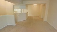 This single-floor space is designed with an open, functional layout that promotes a sense of connectivity between its main areas. The living area is generously sized, finished with wall-to-wall beige carpeting that adds a soft, neutral warmth underfoot. Clean white walls and simple baseboard trim run throughout, contributing to a bright and understated aesthetic.

A defining architectural feature is the half-wall pass-through that separates the kitchen from the living space. This design element maintains visual openness while clearly delineating the two areas. The kitchen, visible through the pass-through, is equipped with white appliances, including a refrigerator and microwave, and features white cabinetry. The curved countertop edge of the pass-through adds a subtle design detail to an otherwise straightforward layout.

Overhead, a structural soffit runs along the ceiling, adding dimension to the space and framing the transition between the kitchen and living areas. Recessed flush-mount ceiling lighting provides practical illumination. The overall ambiance is clean, neutral, and move-in ready, offering a versatile blank canvas well-suited to a variety of furnishing styles and personal tastes.