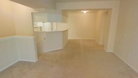 This single-floor space is designed with an open, functional layout that promotes a sense of connectivity between its main areas. The living area is generously sized, finished with wall-to-wall beige carpeting that adds a soft, neutral warmth underfoot. Clean white walls and simple baseboard trim run throughout, contributing to a bright and understated aesthetic.

A defining architectural feature is the half-wall pass-through that separates the kitchen from the living space. This design element maintains visual openness while clearly delineating the two areas. The kitchen, visible through the pass-through, is equipped with white appliances, including a refrigerator and microwave, and features white cabinetry. The curved countertop edge of the pass-through adds a subtle design detail to an otherwise straightforward layout.

Overhead, a structural soffit runs along the ceiling, adding dimension to the space and framing the transition between the kitchen and living areas. Recessed flush-mount ceiling lighting provides practical illumination. The overall ambiance is clean, neutral, and move-in ready, offering a versatile blank canvas well-suited to a variety of furnishing styles and personal tastes.
