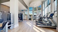 Located at 6050 Roswell Rd in Sandy Springs, GA, this contemporary fitness center is designed to deliver a bright, energizing workout environment. The space is defined by soaring ceilings and an expansive wall of floor-to-ceiling windows that flood the interior with natural light and offer open views of the surrounding Sandy Springs cityscape and greenery. Light hardwood flooring runs throughout, complementing the clean, modern aesthetic of the white and gray walls.

The cardio zone is generously equipped with multiple treadmills, elliptical trainers, stair climbers, stationary bikes, and a rowing machine — all positioned to take full advantage of the sweeping street-level views outside. A dedicated strength training area features a dumbbell rack, weight benches, and a multi-function cable machine, providing options for a full range of resistance workouts. A functional training corner rounds out the facility with TRX suspension straps, a heavy punching bag, and a well-stocked rack of fitness accessories including kettlebells and medicine balls.

Accent lighting from pendant fixtures adds warmth to select areas, while built-in cubby storage and wall-mounted televisions enhance the overall functionality of the space. The layout is thoughtfully organized, allowing users to move fluidly between workout zones in a well-lit, spacious, and motivating environment.