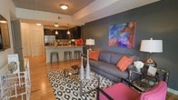 This single-floor residence showcases a thoughtfully designed open-concept layout that seamlessly connects the living area and kitchen. The living space is anchored by a plush gray sofa dressed with vibrant orange accent pillows, positioned atop a bold geometric-patterned area rug in black and white. A striking dark accent wall serves as the backdrop for a vivid, colorful abstract artwork, adding a strong focal point to the room. Warm hardwood flooring runs throughout, lending the space a cohesive and inviting feel. The kitchen is a standout feature, offering dark cabinetry paired with light granite countertops and a peninsula-style breakfast bar lined with industrial metal barstools. Pendant lighting with warm-toned bulbs illuminates the kitchen area, complementing the recessed ceiling lights that brighten the overall space. Stainless steel appliances add a sleek, modern edge to the kitchen. The open sightlines between the living and kitchen areas create a sense of expansive flow, making the layout ideal for both everyday living and entertaining. The overall ambiance is contemporary and vibrant, balancing bold color choices with clean, modern design elements.