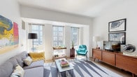 This single-floor residence offers a thoughtfully designed layout that balances contemporary style with everyday functionality. The living area is a standout space, characterized by an abundance of natural light streaming through a bank of large, gridded windows that frame urban views of the surrounding cityscape. White walls and light hardwood flooring create a clean, airy backdrop, while the open layout allows for flexible furnishing arrangements. A bold abstract artwork anchors one wall, adding a vibrant focal point to the room.

The kitchen is efficiently designed with soft grey cabinetry accented by brass hardware, white quartz countertops, and a striking marble-patterned tile backsplash. A central island with bar seating provides additional prep space and casual dining. Full-sized white appliances and generous cabinet storage complete the practical layout, illuminated by a sleek brass pendant fixture overhead.

The bedroom is quietly tucked away, benefiting from large windows that invite natural light and offer outdoor views. A double-door closet provides ample storage. The full bathroom features dark subway tile surrounding a full soaking tub, dark floor tile, and a stacked in-unit washer and dryer conveniently integrated within the space.