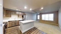 This single-floor space showcases a thoughtfully designed open-concept layout that seamlessly integrates the kitchen and living areas. The kitchen is outfitted with rich dark wood cabinetry, stainless steel appliances including a refrigerator and dishwasher, and a crisp white countertop island that serves as both a workspace and a natural divider between the kitchen and living areas. A stainless steel sink with modern fixtures is positioned beneath recessed and track lighting, ensuring the kitchen is well-lit for both cooking and entertaining. The flooring transitions from durable dark hardwood in the kitchen to plush carpet in the living area, providing both practicality and comfort. The living area is generously sized and bathed in natural light courtesy of large windows along the far wall, creating a bright and welcoming ambiance. Neutral gray walls complement the warm tones of the cabinetry and flooring throughout, lending the space a cohesive and contemporary feel. Recessed lighting is distributed throughout the ceiling, offering ample illumination. The overall design balances functionality and style, making this space well-suited for everyday living and casual entertaining alike.