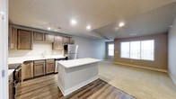 This single-floor space showcases a thoughtfully designed open-concept layout that seamlessly integrates the kitchen and living areas. The kitchen is outfitted with rich dark wood cabinetry, stainless steel appliances including a refrigerator and dishwasher, and a crisp white countertop island that serves as both a workspace and a natural divider between the kitchen and living areas. A stainless steel sink with modern fixtures is positioned beneath recessed and track lighting, ensuring the kitchen is well-lit for both cooking and entertaining. The flooring transitions from durable dark hardwood in the kitchen to plush carpet in the living area, providing both practicality and comfort. The living area is generously sized and bathed in natural light courtesy of large windows along the far wall, creating a bright and welcoming ambiance. Neutral gray walls complement the warm tones of the cabinetry and flooring throughout, lending the space a cohesive and contemporary feel. Recessed lighting is distributed throughout the ceiling, offering ample illumination. The overall design balances functionality and style, making this space well-suited for everyday living and casual entertaining alike.