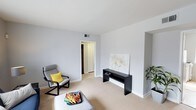 This single-floor space presents a living area defined by its clean, minimalist aesthetic and neutral color palette. The walls are painted in a soft, light gray tone that lends the room an airy, open quality, complemented by white trim and baseboards that frame the space with crisp definition. Underfoot, wall-to-wall beige carpeting adds warmth and contributes to the room's quiet, comfortable atmosphere.

The layout is straightforward and uncluttered. A modern rocking chair in light gray upholstery, accented with a bright yellow throw pillow, occupies one corner, providing a casual seating option. Nearby, a low-profile ottoman serves as additional functional furnishing. A sleek, dark console table is positioned along one wall beneath a canvas print featuring soft floral imagery, adding a subtle decorative focal point to the room. A potted plant near the far wall introduces a natural element to the otherwise simple interior.

A doorway with white trim leads to an adjacent area, suggesting further rooms within the unit. Ceiling-mounted ventilation is visible overhead, and the smooth white ceiling maintains the room's clean, unadorned character. Overall, the space conveys a calm, practical, and well-maintained environment with a modern sensibility.