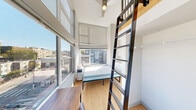Located on the sixth floor at 1815 Bellevue Ave in Seattle's vibrant Capitol Hill neighborhood, Unit 607 is a thoughtfully designed loft-style studio that maximizes vertical space through impressive double-height ceilings. A sleek, dark metal ladder leads up to an open mezzanine level finished with warm wood accents and railing, providing an elevated sleeping or storage area with a ceiling fan overhead.

The main living area features light-toned hardwood flooring and is bathed in abundant natural light from expansive floor-to-ceiling windows on two sides, offering open views of the surrounding Seattle cityscape. A dedicated workspace with a wood desk and chair is positioned to take advantage of the views.

The compact kitchenette is efficiently outfitted with upper and lower white cabinetry, a countertop workspace, a compact refrigerator, and a microwave. The bathroom includes a stand-up shower, toilet, vanity with mirror, and warm-toned flooring, with a wood-paneled ceiling adding a natural design touch.

A walk-in closet area is conveniently located beneath the mezzanine. The space is accented by a bold teal entryway door, adding a pop of color to the otherwise clean, modern white interior palette. This well-appointed urban studio offers an efficient and stylish living solution in the heart of Seattle.
