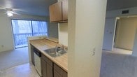 This single-floor residential space offers a practical and functional layout designed for comfortable everyday living. The kitchen serves as a central feature, showcasing warm wood-toned cabinetry both above and below the countertops, providing generous storage throughout. A light-colored laminate countertop runs along the workspace, complemented by a stainless steel single-basin sink and a built-in dishwasher, making the kitchen both efficient and easy to maintain. A textured accent wall adds subtle depth and visual interest to the space. Natural light filters in through a large window dressed with vertical blinds, brightening the kitchen and offering a view to the outside. The open pass-through design connects the kitchen to the adjacent carpeted living area, creating a sense of flow and spaciousness between the two zones. The tile flooring in the kitchen transitions seamlessly into the soft carpeting of the living space, distinguishing functional areas while maintaining a cohesive overall feel. The neutral color palette throughout the unit provides a clean, versatile backdrop suitable for a variety of interior styles. Overall, the space combines practical amenities with an open, airy atmosphere.