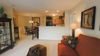 This single-floor residence offers a cohesive and welcoming open-concept layout that seamlessly connects the main living areas. The living room is comfortably appointed with warm-toned seating, decorative accent pillows, and soft carpeting underfoot, creating a relaxed and inviting atmosphere. A half-wall peninsula separates the kitchen from the main living space while maintaining an open sightline throughout. The kitchen features light wood cabinetry, black appliances, and decorative greenery accents atop the upper cabinets, adding a touch of natural charm. Recessed lighting and a flush-mount ceiling fixture provide ample illumination throughout the kitchen area. Adjacent to the kitchen, the dining area accommodates a classic dark wood table with ornate chair detailing, lit by a chandelier overhead that adds a traditional elegance. A framed piece of wall art anchors the dining nook and lends character to the space. The neutral color palette of warm beige tones on the walls and carpet ties each area together harmoniously. Textured ceilings and thoughtful lighting throughout contribute to the overall warmth and livability of this well-designed single-level home.