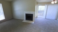 This single-floor residence offers a comfortable and functional layout designed for everyday living. The living area serves as the heart of the space, anchored by a classic fireplace framed in a light marble-style tile surround, adding a refined focal point to the room. Soft, wall-to-wall carpeting extends throughout, contributing to a warm and inviting atmosphere underfoot. The room is bathed in natural light, which streams in through a sliding glass door that leads directly to an outdoor balcony. The balcony, enclosed by a wooden railing, provides a pleasant outdoor extension of the living space with views of surrounding greenery. The neutral color palette of the walls and flooring creates a versatile backdrop, allowing the space to be easily personalized. A textured ceiling adds a subtle architectural element overhead. The open flow between the living area and the balcony enhances the sense of space and connectivity to the outdoors, making this residence a practical and comfortable place to call home.