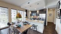 This single-floor space showcases a seamlessly integrated kitchen and dining area designed with a contemporary aesthetic. The kitchen is anchored by a large peninsula with striking granite countertops, offering bar seating and serving as a natural divider between the cooking and dining zones. Dark, sophisticated cabinetry contrasts beautifully with a warm brick tile backsplash, while stainless steel appliances — including a French door refrigerator, microwave, and range — add a polished, professional feel. Pendant lighting in cylindrical glass fixtures hangs above the peninsula, providing focused illumination and a refined decorative touch. Rich, dark hardwood flooring flows throughout the open layout, grounding the space with warmth and continuity. The dining area comfortably accommodates a full-sized wooden table and benefits from generous natural light streaming through large windows dressed with horizontal blinds. The tray ceiling detail above the kitchen zone adds architectural depth and visual interest. Overall, the space delivers a cohesive, upscale ambiance that balances functionality with modern design sensibility.