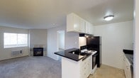 Located at 1845 N Broadway in Escondido, this single-story unit offers a functional and cohesive layout with an open connection between the kitchen and living area. The kitchen is outfitted with white cabinetry extending to the ceiling, providing ample storage, and is anchored by a peninsula with dark granite countertops. Black appliances, including an electric range and refrigerator, complement the contrasting color scheme. Light tile flooring runs through the kitchen, transitioning to carpet in the adjoining living space. The living area features a tiled fireplace with a classic surround, adding a focal point to the room. A textured ceiling is consistent throughout the space. A doorway visible from the main living area leads to an additional room, suggesting a practical and connected floor plan. The overall palette of white, grey, and black creates a clean, neutral aesthetic throughout the unit.