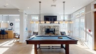 This single-floor community clubhouse in East Providence, Rhode Island, is designed to offer a range of social and recreational amenities within a bright, welcoming environment. The interior is finished in soft gray and white tones throughout, with recessed lighting, crown molding, and neutral carpeting that contribute to a clean, cohesive aesthetic.

The expansive main lounge serves as the heart of the space, furnished with comfortable sofas and upholstered armchairs arranged around low coffee tables. Large windows along the perimeter flood the area with natural light, while structural columns define zones without closing off the open floor plan. A dining counter with seating provides an additional gathering spot within this central area.

The game room is a standout feature, centered around a full-size pool table illuminated by dual pendant light fixtures. A stone fireplace with a wall-mounted television above anchors one wall, flanked by windows that bring in additional daylight. French doors nearby provide access to the outdoors.

A compact kitchenette offers light maple cabinetry, quartz countertops, a sink, microwave, coffee maker, and ice maker for resident convenience. A small conference room accommodates intimate meetings with a round table and ergonomic seating. A casual activity nook rounds out the space, featuring bistro-height seating, a chess set, and local artwork.