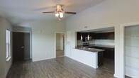 This single-story space showcases an open-concept layout that seamlessly connects the living area and kitchen. The living area features soaring vaulted ceilings accentuated by a ceiling fan with integrated lighting, contributing to an airy and spacious atmosphere. Rich, dark hardwood-style flooring runs throughout, providing a warm and cohesive aesthetic.

The kitchen is a standout feature, designed with dark espresso cabinetry that contrasts sharply against crisp white walls. Granite countertops extend across the workspace and wrap around a peninsula that separates the kitchen from the living area, offering both additional prep space and a casual seating option. Stainless steel appliances, including a range and microwave, are integrated into the cabinetry for a streamlined look. Pendant lighting hangs above the peninsula, adding focused illumination and a modern design touch.

A hallway visible from the main living area leads to additional rooms, suggesting a thoughtful and functional floor plan. The neutral wall tones throughout complement the darker finishes, creating a balanced and contemporary interior that is both practical and visually appealing.
