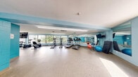 Located at 545–565 Belmont Ave W in Kitchener, Ontario, this fitness facility offers a comprehensive, single-floor exercise space designed to support a wide range of workout routines. The room is finished with durable speckled flooring throughout, and the walls are painted in a fresh light blue palette accented by bold teal feature walls, creating an energetic yet clean atmosphere.

The space is divided into distinct functional zones. A dedicated cardio area lines one side of the room, equipped with treadmills, elliptical trainers, and a stationary bike, all positioned in front of large floor-to-ceiling windows that bring in generous natural light and views of the surrounding greenery. Full-length mirrors run along the walls, enhancing the sense of openness and allowing users to monitor their form.

A strength training zone houses a multi-station cable machine and rowing machines, providing options for resistance and full-body workouts. An additional open-concept area with wall-mounted mirrors and a mounted flat-screen TV offers flexible space well-suited for stretching, group classes, or functional training. Bright overhead fluorescent lighting ensures consistent illumination throughout all areas.

The facility is well-ventilated with visible ceiling vents and is thoughtfully appointed with practical amenities including a wall-mounted TV, a portable fan, and conveniently placed waste receptacles, making it a functional and welcoming fitness environment.