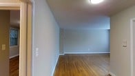 This single-floor space presents a clean and straightforward layout designed for practical, everyday living. The interior is characterized by smooth, neutral-toned walls painted in a soft gray that lend the space a calm and understated ambiance. Warm-toned hardwood flooring runs throughout, adding natural richness and a sense of continuity to the open area. The flooring displays a classic plank pattern with a polished finish that reflects the overhead lighting, contributing to the overall brightness of the space. Crisp white baseboards provide a clean contrast against the walls and flooring, adding a neat, finished look to the perimeter of each room. Ceiling-mounted flush lighting provides even illumination across the space. A doorway is visible along one wall, hinting at additional rooms or access points within the layout. The walls are free of ornamentation, creating a blank canvas that allows for flexible interior styling and personalization. Overall, this space offers a modest yet well-maintained environment with a neutral palette and classic hardwood flooring that form a solid foundation for comfortable, functional use.