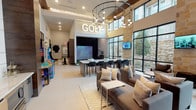 This contemporary amenity lounge is designed with entertainment and social engagement at its core. Soaring double-height ceilings draw the eye upward, accentuated by a striking circular chandelier with pendant bulbs that casts warm, ambient light across the open-plan space. Large floor-to-ceiling windows framed in sleek black steel flood the interior with natural light while offering views of a natural stone-accented exterior courtyard, reinforcing a connection to the outdoors.

The centerpiece of the space is a dedicated golf simulator bay, prominently marked with an illuminated marquee "GOLF" sign. Adjacent to the simulator, a long marble-topped bar counter with woven leather barstools provides an ideal perch for spectating and socializing. Warm wood-paneled ceilings above this zone add a refined, inviting contrast to the otherwise light and airy palette.

A comfortable lounge area anchors the main gathering space, featuring plush sofas and accent chairs arranged around a coffee table on a patterned rug. A large wall-mounted flat-screen TV with built-in cabinetry and wet bar serves as the entertainment focal point. Classic arcade game cabinets add a playful, nostalgic touch to the atmosphere.

The entrance is defined by elegant glass-paneled double doors with black steel framing and is complemented by lush oversized indoor planters, creating a welcoming transition into this vibrant, well-appointed social hub.