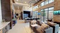This contemporary amenity lounge is designed with entertainment and social engagement at its core. Soaring double-height ceilings draw the eye upward, accentuated by a striking circular chandelier with pendant bulbs that casts warm, ambient light across the open-plan space. Large floor-to-ceiling windows framed in sleek black steel flood the interior with natural light while offering views of a natural stone-accented exterior courtyard, reinforcing a connection to the outdoors.

The centerpiece of the space is a dedicated golf simulator bay, prominently marked with an illuminated marquee "GOLF" sign. Adjacent to the simulator, a long marble-topped bar counter with woven leather barstools provides an ideal perch for spectating and socializing. Warm wood-paneled ceilings above this zone add a refined, inviting contrast to the otherwise light and airy palette.

A comfortable lounge area anchors the main gathering space, featuring plush sofas and accent chairs arranged around a coffee table on a patterned rug. A large wall-mounted flat-screen TV with built-in cabinetry and wet bar serves as the entertainment focal point. Classic arcade game cabinets add a playful, nostalgic touch to the atmosphere.

The entrance is defined by elegant glass-paneled double doors with black steel framing and is complemented by lush oversized indoor planters, creating a welcoming transition into this vibrant, well-appointed social hub.