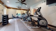 Located at Hanover Crossings in Mechanicsville, Virginia, this fitness center offers a comprehensive workout facility designed to support a wide range of exercise routines. The space is thoughtfully laid out across a single floor, with distinct areas dedicated to cardio training, strength work, and functional fitness.

The cardio section is well-stocked with multiple treadmills — including Spirit and Matrix branded models — along with Matrix elliptical trainers and a stationary bike, providing ample options for aerobic exercise. Ceiling fans throughout the space help maintain airflow and comfort during workouts. Recessed lighting and roller shade-equipped windows allow natural light to filter in while giving users control over glare.

The strength and functional training area features a multi-station cable machine, an adjustable weight bench, a full rack of dumbbells, kettlebells, and a variety of medicine balls and exercise balls neatly organized on a dedicated rack. A large wall mirror enhances the sense of space and allows users to monitor their form during exercise.

Warm wood-toned crown molding with orange accent trim adds a polished, upscale aesthetic to the room, complemented by neutral-toned carpet flooring with stripe detailing. A wall-mounted television is available for entertainment. Direct exterior door access provides convenient entry and exit, contributing to the overall functionality of this well-appointed fitness facility.