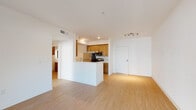 This well-appointed single-floor residence offers a clean, contemporary design with an open-concept layout that seamlessly connects the living area and kitchen. Light wood-tone flooring flows throughout the main living spaces, creating a warm and cohesive aesthetic. The kitchen is anchored by a central peninsula with a built-in sink, providing both workspace and a casual dining or entertaining area. Warm wood cabinetry pairs with stainless steel appliances — including a gas range, over-the-range microwave, refrigerator, and dishwasher — set against a modern backsplash. The open living area is bright and spacious, with natural light entering through a glass door that leads to an outdoor area. The two bedrooms feature plush carpeting, clean white walls, and generous closet access, offering a comfortable and quiet retreat. Both bathrooms are finished with large-format tile flooring, wood-toned cabinetry, white quartz countertops, and a tub-shower combination enclosed by sliding glass doors with striking marble-effect tile surround. The overall design emphasizes simplicity and functionality, with a neutral color palette and modern finishes that create a bright, airy atmosphere throughout.