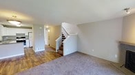 This two-story home offers a functional and straightforward layout across two levels, providing comfortable living space for its occupants. The main level features a combination of wood-look hard flooring and neutral-toned carpet, creating a clear distinction between different areas of the home. A central staircase with a white-painted banister connects the two floors, serving as a natural focal point within the open living area.

The main level living space is finished with soft gray walls and standard ceiling heights, giving the area a clean, understated appearance. A hallway leads toward the kitchen, where a refrigerator is visible, indicating a practical galley-style or compact kitchen layout. Floor vents throughout the main level suggest a forced-air climate control system. The transition between the hard flooring near the entryway and kitchen and the carpeted living area helps define the functional zones of the space.

With two bedrooms and one and a half bathrooms spread across both floors, the home provides a sensible separation between living and sleeping areas. The overall design is neutral and move-in ready, with a simple, clean aesthetic throughout.