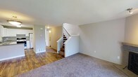 This two-story home offers a functional and straightforward layout across two levels, providing comfortable living space for its occupants. The main level features a combination of wood-look hard flooring and neutral-toned carpet, creating a clear distinction between different areas of the home. A central staircase with a white-painted banister connects the two floors, serving as a natural focal point within the open living area.

The main level living space is finished with soft gray walls and standard ceiling heights, giving the area a clean, understated appearance. A hallway leads toward the kitchen, where a refrigerator is visible, indicating a practical galley-style or compact kitchen layout. Floor vents throughout the main level suggest a forced-air climate control system. The transition between the hard flooring near the entryway and kitchen and the carpeted living area helps define the functional zones of the space.

With two bedrooms and one and a half bathrooms spread across both floors, the home provides a sensible separation between living and sleeping areas. The overall design is neutral and move-in ready, with a simple, clean aesthetic throughout.
