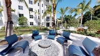 This outdoor amenity space offers a thoughtfully designed retreat defined by tropical landscaping and contemporary furnishings. At the heart of the patio sits a round, bowl-shaped fire pit surrounded by smooth river rock and encircled by sleek, chevron-patterned lounge chairs in a bold blue hue, creating an inviting gathering focal point. The pavers underfoot are light-toned and neatly arranged, contributing to the clean, modern aesthetic of the space.

Mature palm trees rise throughout the area, providing natural shade and reinforcing the tropical ambiance. Lush greenery and manicured plantings border the space, adding depth and color. In the background, a secondary seating area is visible, featuring white lounge chairs beneath a shade sail, offering an additional spot to relax. The entire outdoor space is set against a bright white multi-story building with balconies and large windows, which frames the area and adds an architectural backdrop. Abundant natural sunlight and the surrounding tropical foliage create a resort-like atmosphere ideal for outdoor relaxation and social gatherings.