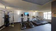 This fitness center offers a practical and well-organized space dedicated to cardiovascular and strength training. The room features dark, durable flooring suited for high-traffic exercise use, complemented by neutral-toned walls that create a clean, focused environment. The textured ceiling adds acoustic character to the space while track lighting ensures the area is well-lit throughout.

The cardio section is a highlight of the facility, equipped with multiple treadmills and elliptical machines positioned to maximize use of the available floor space. A wall-mounted flat-screen television is centrally positioned, allowing exercisers to stay entertained or informed during their workouts. Wall sconces provide additional ambient lighting along the perimeter.

A cable resistance machine is also present, offering users versatile strength training options alongside the cardio equipment. The thoughtful arrangement of machines allows for comfortable movement between stations without congestion.

Overall, the fitness center presents a clean, functional, and fully equipped environment suitable for daily exercise routines, making it a convenient and practical amenity for its users.