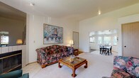 This single-floor residence offers a comfortable and functional layout designed for easy living. The spacious living room serves as the heart of the home, featuring light carpet flooring, a bold floral sofa, a glass-top wood coffee table, and a framed cityscape painting as a focal point on the wall. A tiled fireplace with a brass-trimmed surround anchors one end of the room, complemented by a large window with vertical blinds that fills the space with natural light. The open layout connects seamlessly to the dining area, which is brightened by a prominent skylight and track lighting overhead, along with windows offering outdoor views. A tile-top dining table with classic black Windsor chairs provides an inviting setting for meals. The kitchen is well-equipped with ample wood-trimmed white cabinetry, generous counter space, and a full suite of appliances including a refrigerator, electric range, and dishwasher. Two carpeted bedrooms offer restful retreats, each with closet access and windows. The primary bedroom includes an ensuite bathroom, while the bathroom features a vanity with a integrated sink, medicine cabinet, and mirror. Throughout the home, neutral tones and plush carpet create a warm, cohesive ambiance.