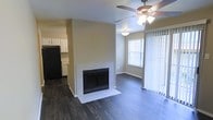 This single-floor residence offers a welcoming and functional layout designed for comfortable everyday living. The living area serves as the heart of the home, anchored by a classic tiled fireplace with a clean white surround that adds warmth and character to the space. Dark hardwood-style flooring runs throughout, providing a rich contrast against the neutral, light-toned walls and lending a sense of depth and sophistication to the interior. A ceiling fan with an integrated light fixture is centrally positioned overhead, ensuring year-round comfort and adequate illumination. Natural light filters in through a large window and a sliding glass door fitted with vertical blinds, brightening the open living and dining area. The layout flows seamlessly into the kitchen, which is equipped with white cabinetry offering ample storage, and a black refrigerator that adds a modern touch. The dining area sits adjacent to the living space, benefiting from the natural light provided by the window. The overall ambiance is clean, neutral, and move-in ready, combining practical features with a simple, understated aesthetic that provides a versatile backdrop for a variety of interior styles.