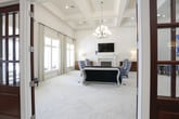 This single-floor space showcases a refined interior design that seamlessly blends classical architectural elements with contemporary furnishings. The layout is anchored by a spacious living area featuring soaring ceilings adorned with coffered detailing and a statement chandelier, creating an immediate sense of grandeur upon entry.

The living area centers around a white-paneled fireplace wall, which extends nearly to the ceiling and serves as a dramatic focal point. A wall-mounted flat-screen television is positioned above the fireplace mantle, flanked by decorative molding panels. Bold blue-and-white animal-print upholstered wingback chairs are arranged around a glass-top coffee table and a cylindrical metallic side table, adding a vibrant contrast against the pale herringbone-patterned flooring.

Natural light filters through multiple windows dressed in patterned drapery, illuminating the crisp white walls and light-toned floors. A rich walnut credenza provides warm contrast alongside the cool palette of the room.

An adjacent seating nook features a dark wood table surrounded by blue-and-white striped chairs, with curated artwork — including a large abstract blue canvas and a grid of gold geometric panels — decorating the tall walls. The grand entryway, with its ornate door and high ceilings, adds a welcoming, sophisticated ambiance to the overall space.