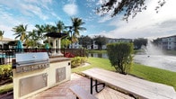 This single-story outdoor space offers a well-appointed entertaining area designed for leisure and al fresco dining. The focal point is a built-in outdoor kitchen station, complete with a stainless steel grill and cabinetry finished in a neutral stucco exterior with a granite-style countertop — a practical and durable setup for outdoor cooking. Adjacent to the grill station, a classic wooden picnic table provides comfortable communal seating on a warm terra cotta tiled patio surface.

The space is framed by abundant tropical landscaping, including mature palm trees, shade trees, and manicured shrubs with vibrant red foliage accents. A charming pergola with white columns adds a decorative architectural element nearby, enhancing the resort-like ambiance.

The patio opens directly to a tranquil waterfront view, with a calm pond or lake reflecting the surrounding greenery and sky. Well-maintained open lawn areas border the water's edge, contributing to an expansive, relaxed atmosphere. The overall setting evokes a warm, Florida-inspired outdoor lifestyle with a balance of functional amenities and natural beauty.