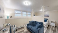Located at 364 Foster Dr in Willimantic, Connecticut, this efficiently designed one-bedroom, one-bathroom unit offers a thoughtfully organized layout across a single floor. The main living area features smooth hardwood-style flooring and warm neutral-toned walls accented by crisp crown molding. Decorative columns subtly define the transition between the living and sleeping areas, creating a sense of structure within the open floor plan. Multiple windows along one wall allow natural light to filter in, brightening the space throughout the day. A built-in wall shelf adds a functional architectural detail, providing display space above the living area. Baseboard heating runs along the perimeter, complemented by a wall-mounted air conditioning unit for year-round climate control. The bedroom area flows seamlessly from the living space and includes two closets for ample storage. The full bathroom is finished with light tile flooring, a bathtub with a curtained shower surround, grab bar, and neutral beige walls. A kitchenette area is visible toward the rear of the unit, equipped with cabinetry and a refrigerator. The overall aesthetic is clean, bright, and functional, making this a practical and comfortable living space.