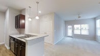 This single-floor residence offers a thoughtfully designed open-concept layout that seamlessly connects the kitchen and living areas. The kitchen is appointed with rich dark cabinetry, a granite-style countertop, and a built-in sink, complemented by stylish pendant lighting that adds a warm, modern touch. The kitchen flows naturally into the living space, which is finished with plush carpeting that adds comfort underfoot. A set of double closet doors provides convenient storage, while a ceiling fan overhead ensures year-round comfort. Natural light fills the living area through a generous window and glass-paneled door, creating a bright and welcoming ambiance. The neutral color palette of soft whites and warm grays throughout the space lends a clean, contemporary feel that is both versatile and inviting. The overall design balances functionality with aesthetic appeal, making it a comfortable and practical living environment. The open sightlines and uncluttered layout enhance the sense of space, while the considered use of lighting — both natural and artificial — ensures every corner of the home feels well-lit and airy.