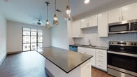 Located at 720 Airport Blvd in Austin, Texas, this single-floor residence offers a thoughtfully designed open-concept layout with contemporary finishes throughout. The kitchen serves as the centerpiece of the living area, featuring crisp white shaker cabinetry, a herringbone-patterned tile backsplash, dark granite countertops, and a central island with pendant lighting in brass-toned fixtures. Stainless steel appliances — including a refrigerator, range, and over-the-range microwave — complete the kitchen's modern, functional aesthetic. Hardwood-style flooring runs throughout the main living areas, adding warmth and continuity to the space. The living area extends toward large floor-to-ceiling windows framed in matte black, allowing abundant natural light and providing views of the surrounding community, including a pool area. The bedroom continues the bright, airy feel with plush carpeting and an expansive grid-style window that further enhances natural light. A ceiling fan is present in both the living and bedroom areas. The bathroom features hardwood-style flooring, a vanity with cabinetry, and a walk-in shower, complemented by clean white walls and practical storage. Recessed lighting throughout the unit ensures consistent illumination across all areas.