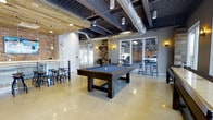 Baypointe's amenity lounge showcases a compelling industrial-chic aesthetic across a single open floor. The space is defined by polished concrete floors that reflect light throughout the room, creating a bright and inviting atmosphere. Exposed ceiling beams with reclaimed wood paneling contrast beautifully against visible ductwork and conduit, lending the space an urban, loft-inspired character.

At the center of the room, a full-size pool table with a blue felt surface serves as the focal point for social gatherings. To one side, a bar-height counter with black industrial-style stools provides casual seating, ideal for socializing or working. Pendant lighting with warm-toned Edison-style bulbs adds a sophisticated layer to the overhead lighting scheme, complementing the wall sconces positioned throughout.

Exposed brick accents and a reclaimed wood feature wall introduce warmth and texture, balancing the cooler tones of the polished floors and gray walls. Large glass doors and windows along the perimeter fill the space with natural light and offer views to the exterior. The overall ambiance is modern yet approachable, designed to encourage community interaction and leisure.