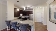 This single-floor residence offers a cohesive blend of modern finishes and warm, inviting design. The open-concept layout connects the kitchen and living area seamlessly, creating a functional and welcoming atmosphere. The kitchen is a standout feature, showcasing rich dark espresso cabinetry, granite countertops, a white subway tile backsplash, and a full suite of stainless steel appliances including a refrigerator, range with hood, and microwave. A large kitchen island with an integrated sink and granite surface doubles as a casual dining bar, complemented by tufted upholstered barstools. Track lighting and pendant fixtures provide ample illumination throughout. The living area features warm hardwood-style flooring, a ceiling fan, and a relaxed, open feel that flows naturally from the kitchen. The bedroom is a serene retreat with plush carpeting, a window with outdoor views, and tasteful neutral tones that create a calm ambiance. The bathroom is equipped with dark cabinetry, granite countertops, accessibility-friendly grab bars, and wood-look tile flooring. A louvered closet door adds a classic touch. Throughout the residence, consistent use of granite, dark wood cabinetry, and neutral walls creates a unified and polished aesthetic.