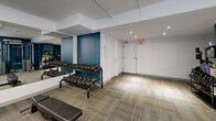 Located at 1231 Richmond St in London, Ontario, this fitness facility offers a thoughtfully organized workout space designed to accommodate a variety of exercise routines. The interior features a clean, modern aesthetic with bold teal accent walls contrasting against crisp white surfaces, all tied together by neutral, textured carpet flooring that runs throughout the main workout area.

The main floor is home to a comprehensive selection of fitness equipment, including a treadmill, stationary spin bike, and a multi-function cable machine for strength training. A rack of dumbbells in graduated weights lines one wall, complemented by adjustable workout benches positioned to allow for a range of exercises. Stability balls in multiple sizes are neatly mounted on the wall, adding versatility for core and balance training. A wall-mounted flat-screen television provides entertainment during workouts.

Large mirrors span one full wall, visually expanding the space and allowing users to monitor their form. Recessed and flush-mounted ceiling lights ensure the area is well-lit, while a window brings in a measure of natural light. The facility also includes an accessible bathroom, rounding out the practical amenities. The space is well-suited for individual fitness routines, offering a focused and functional environment for residents or users seeking a convenient, all-in-one workout facility.