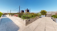 This expansive rooftop terrace offers a well-appointed outdoor retreat above the city skyline. The space is thoughtfully divided into distinct functional zones, each designed for comfort and entertaining. Large raised concrete planters filled with drought-tolerant agaves, ornamental grasses, and colorful flowering plants serve as natural dividers throughout the terrace, adding vibrant greenery against the urban backdrop.

A dedicated dining area features a rustic wood communal table with red metal chairs, positioned alongside a built-in outdoor kitchen equipped with a stainless steel grill — ideal for al fresco gatherings. String lights crisscross overhead, lending the space a warm ambiance after dark.

A separate lounge zone provides a more intimate setting, centered around a round fire pit surrounded by plush, cushioned wicker sofas and accent chairs. A wall-mounted screen adds a versatile entertainment element to this seating area.

Manicured hedgerows line portions of the perimeter, offering both privacy and a refined aesthetic, while open sightlines deliver panoramic views of the surrounding city skyline and distant hills. The entire terrace is finished with large-format porcelain tile pavers, lending a clean, contemporary feel to the overall design.